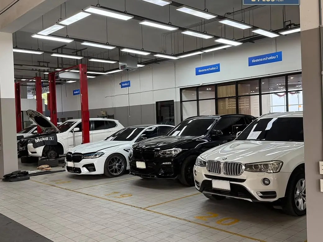 รถ BMW หลายคันจอดเรียงกันในช่องจอดที่มีหลังคาของอู่ P-Plus BM Service มีป้ายกำกับช่องรถระหว่างซ่อม