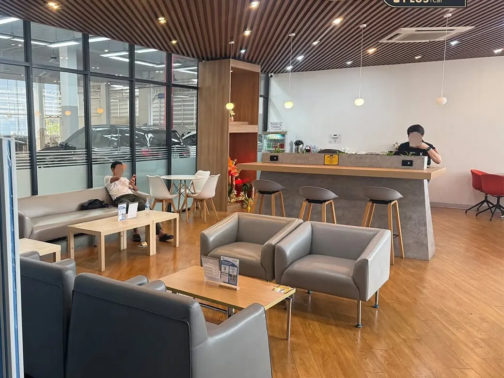 ห้องรับรองลูกค้าของ P-Plus BM Service มีโซฟา โต๊ะไม้ และเคาน์เตอร์กาแฟ มองเห็นพื้นที่ซ่อมผ่านผนังกระจก