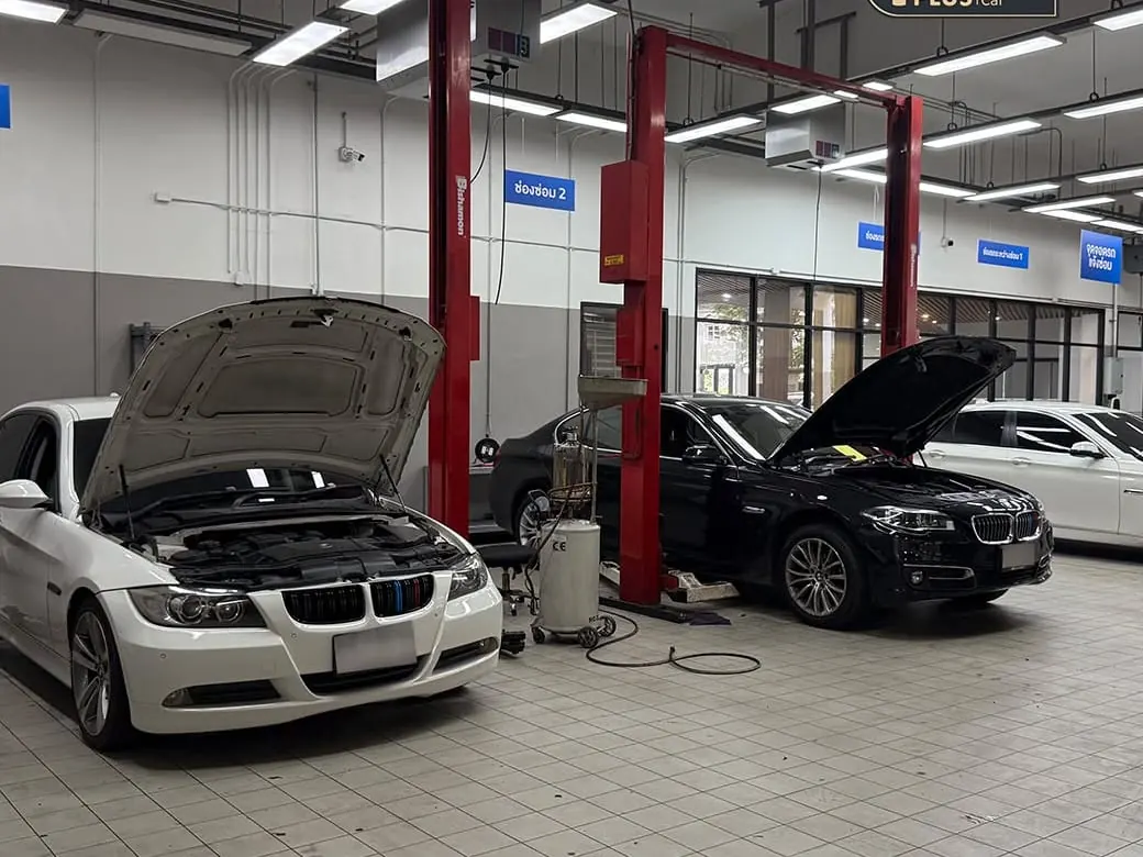 รถ BMW สองคันจอดเปิดฝากระโปรงอยู่ในช่องซ่อมของอู่ P-Plus BM Service พร้อมถังถ่ายน้ำมันเครื่องระหว่างเข้าเช็คระยะ